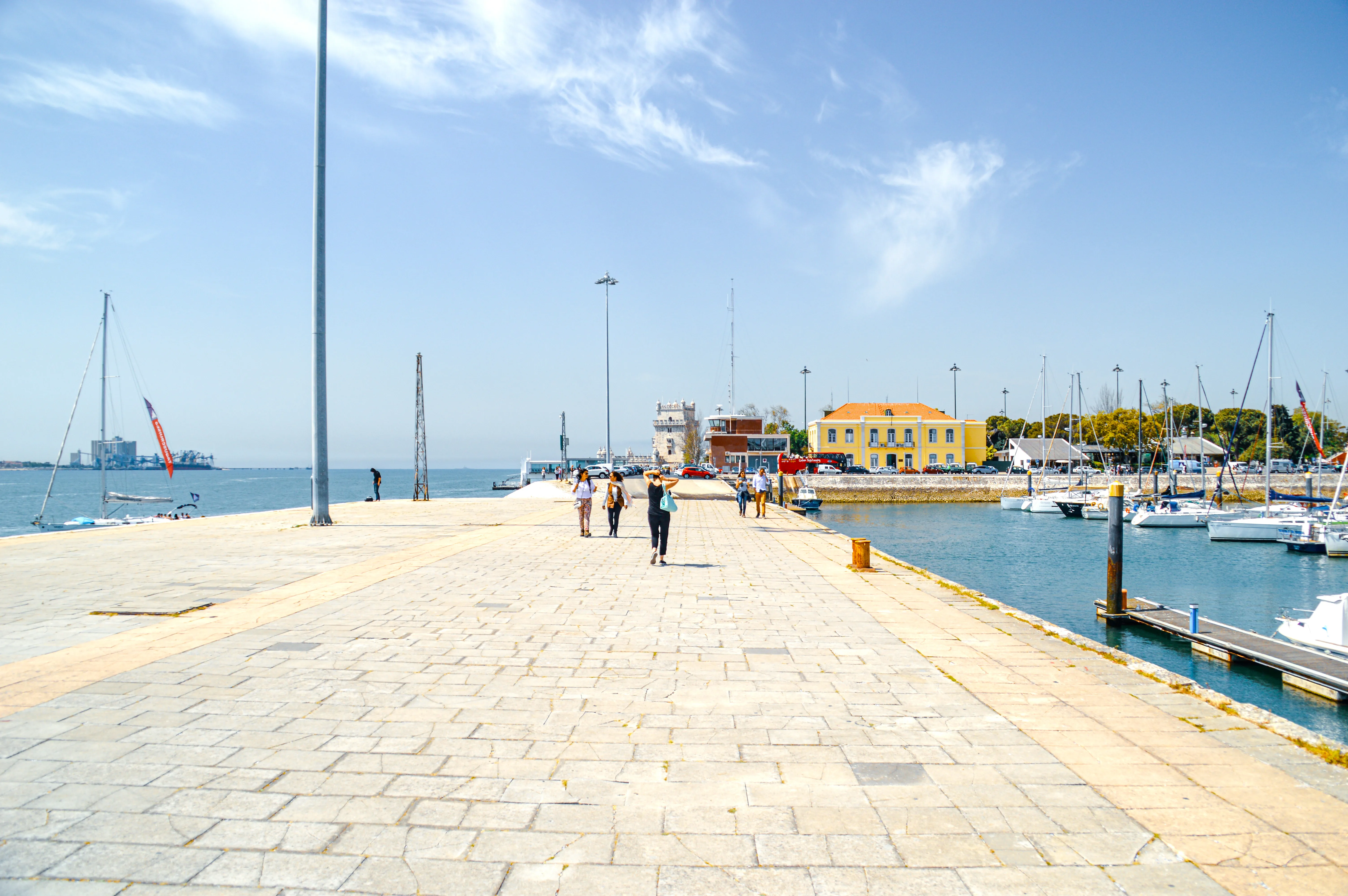 People walking on a stone promenade next to a marina filled with sailboats under a clear blue sky.